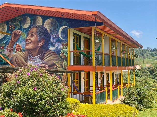 CREATOR gd-jpeg v10 using IJG JPEG v62 default quality - Alquiler de Fincas en el Quindio, Hoteles en el Eje Cafetero Two-story colorful building with yellow railings and green doors; large mural of a smiling Indigenous woman painting against a starry, moonlit background.